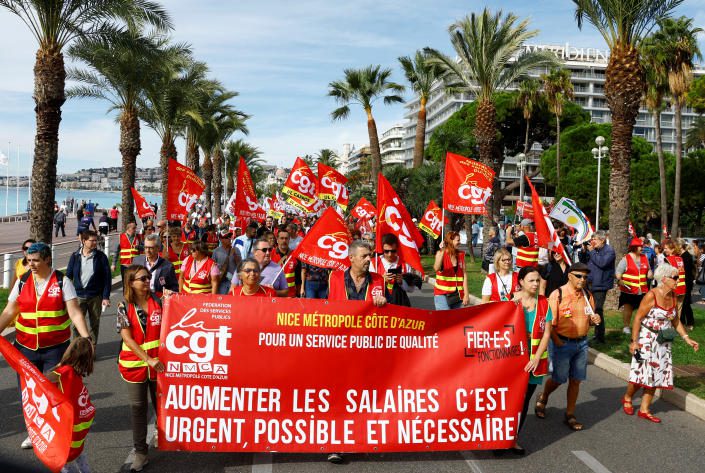 I lavoratori sindacali francesi camminano lungo una strada fiancheggiata da palme, sventolando bandiere scarlatte e uno striscione abbinato.