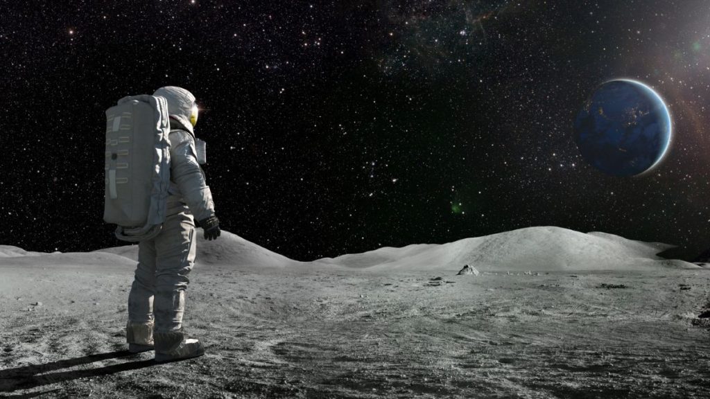 A lone astronaut standing facing away from the camera dressed in full space suit with backpack, stands still looking towards a distant planet Earth.