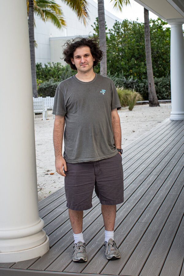 Sam Bankman-Fried stands outdoors in front of a large, white column on a porch near palm trees.